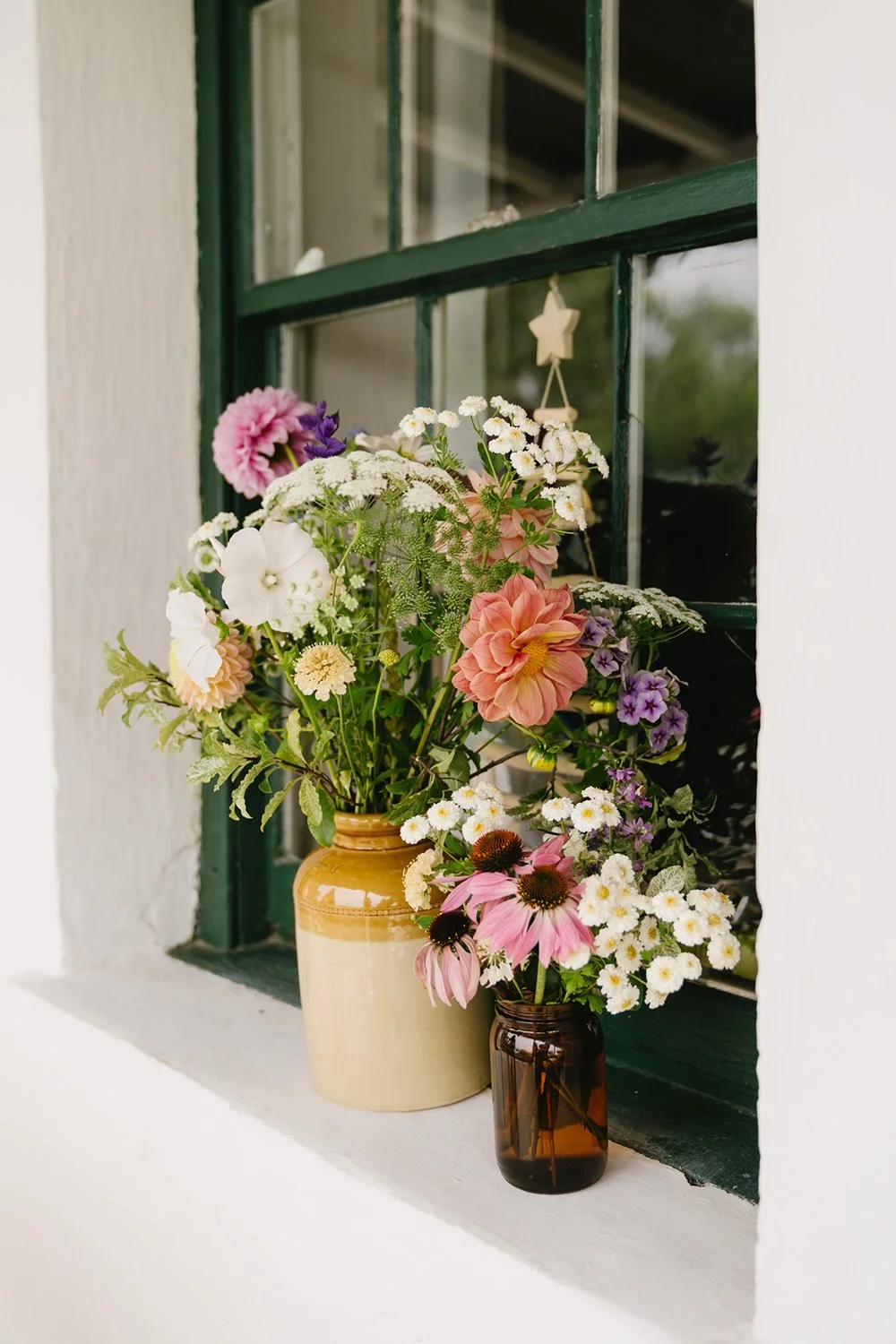 A window with green panes, containing two vases filled with a variety of colorful flowers on the windowsill.