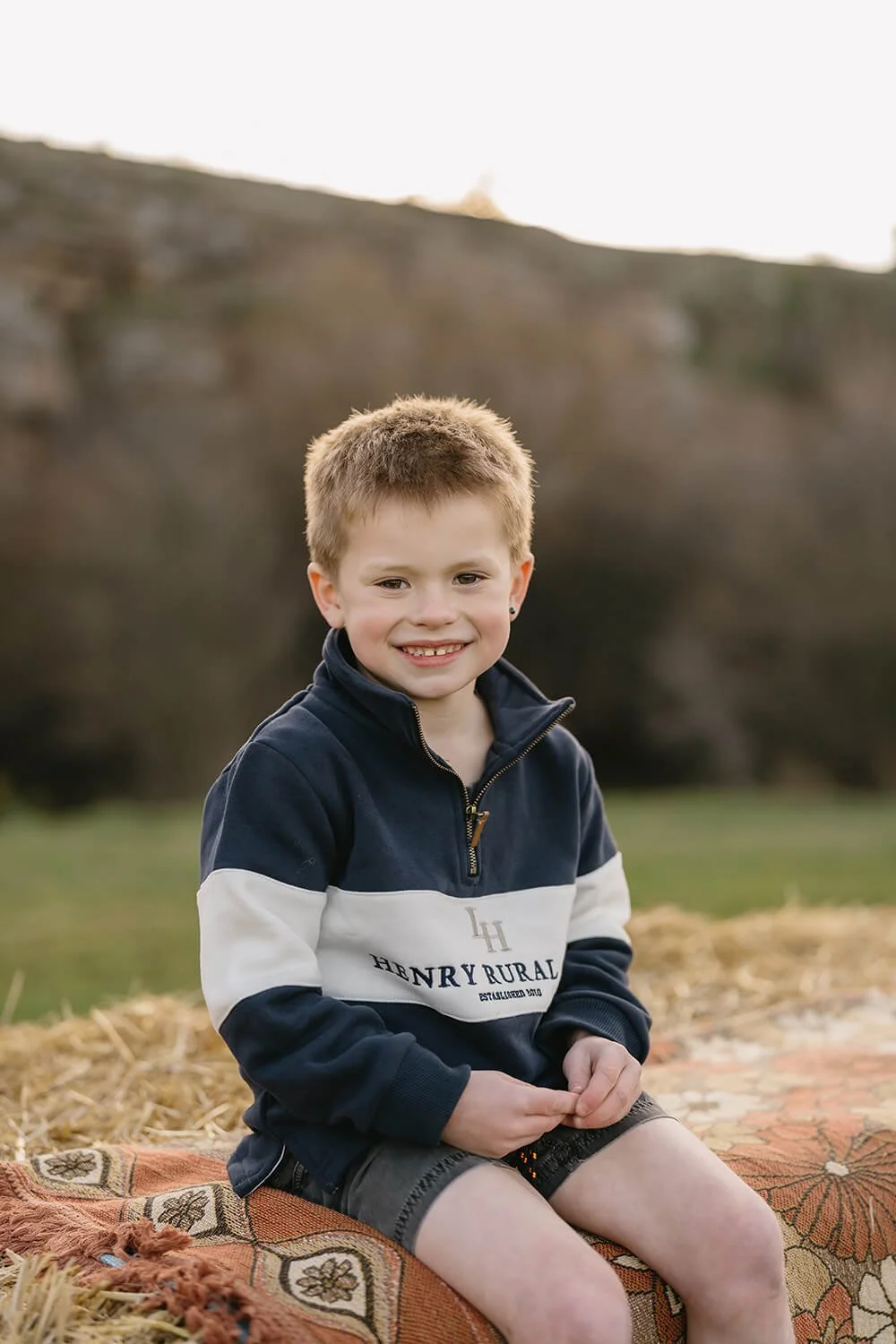 A young boy with short light brown hair and a smile, sitting on a blanket outdoors with a grassy field and a rocky hillside in the background, during late afternoon or early evening.
