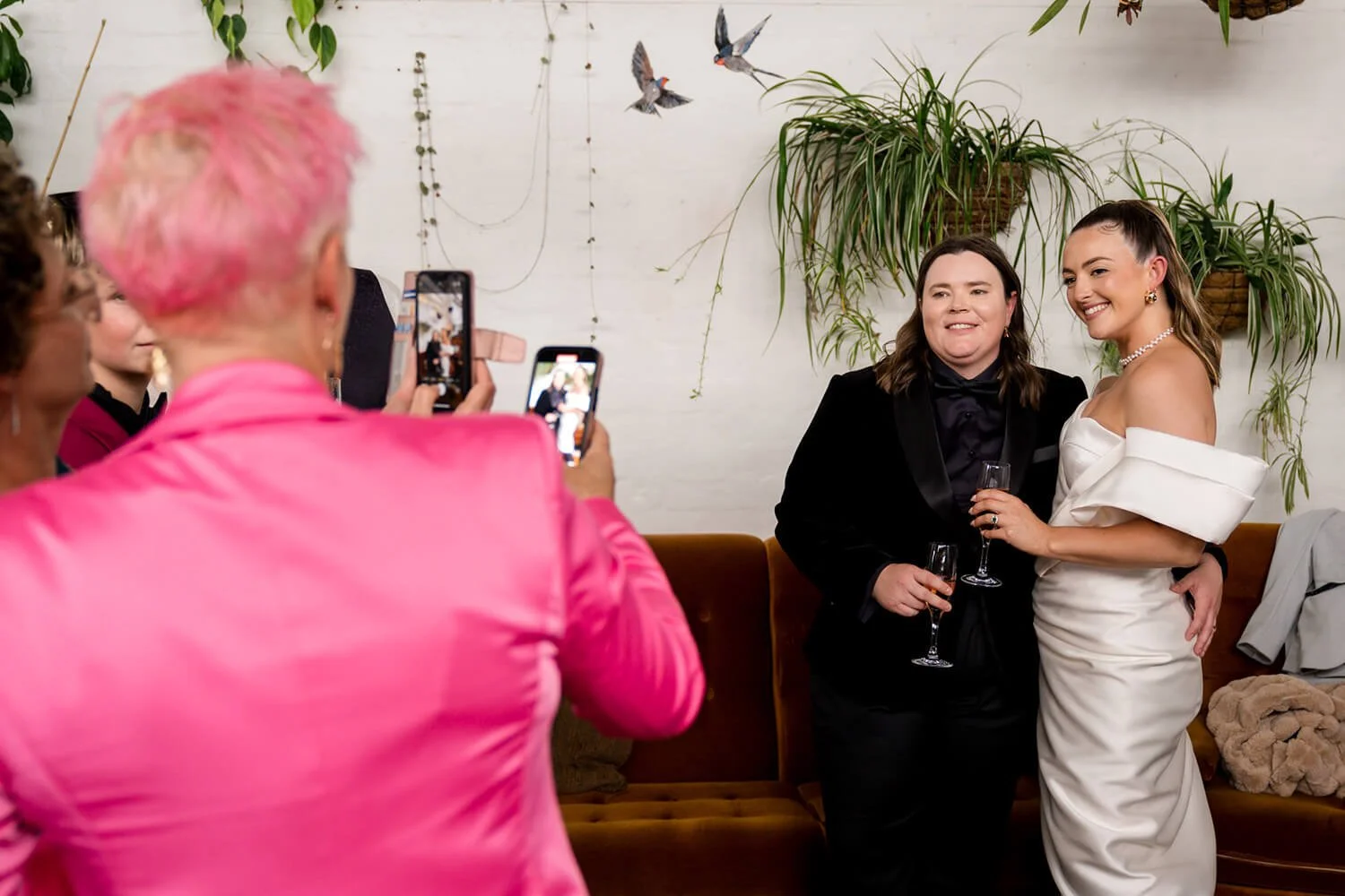 Two women, one in a black tuxedo and the other in a white off-shoulder dress, pose together for photographs with glasses of champagne in a decorated indoor setting with plants and wall art shaped like birds.