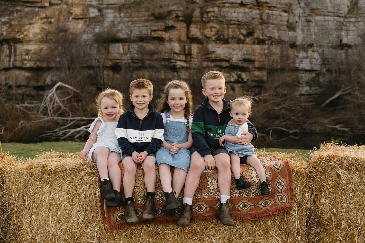 A group of six children sitting on a hay bale outdoors, with a rocky cliff in the background during daytime.