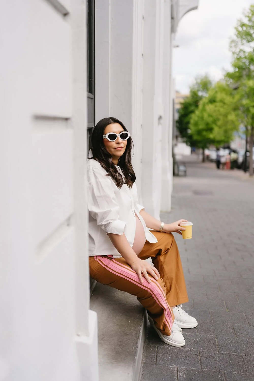 Pregnant woman sitting on a city sidewalk, wearing white shirt, brown pants with pink stripes, white sneakers, black sunglasses, holding a coffee to go, leaning against white building wall, trees and street in the background.