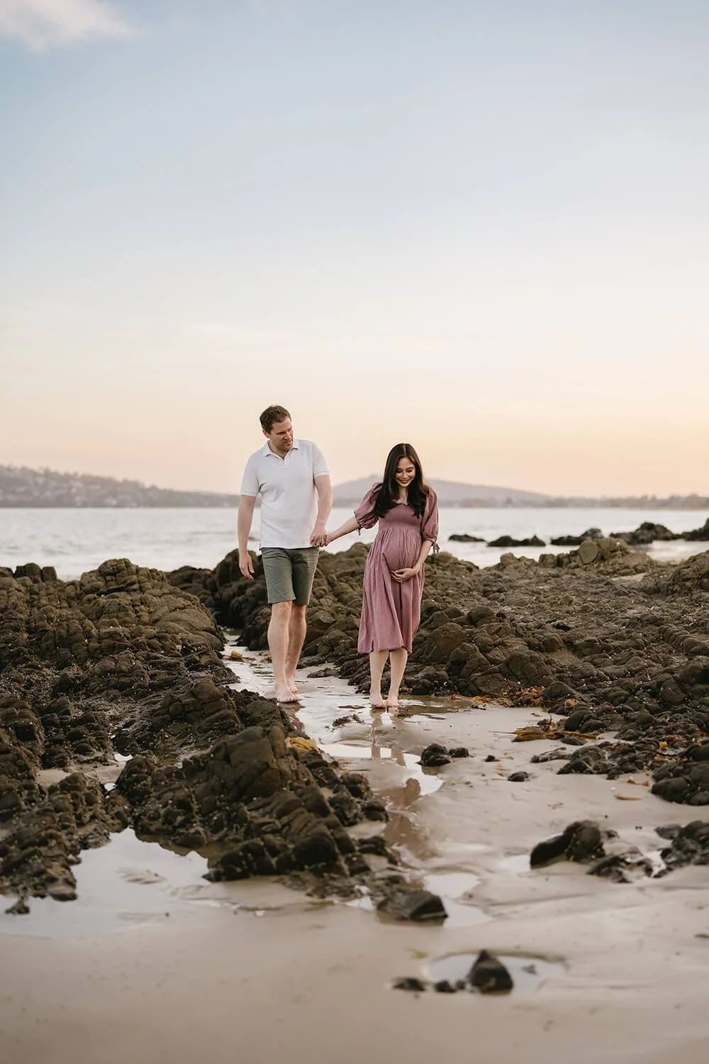A pregnant woman in a mauve dress walking on rocks at the beach with a man in a white shirt and shorts holding her hand during sunset.