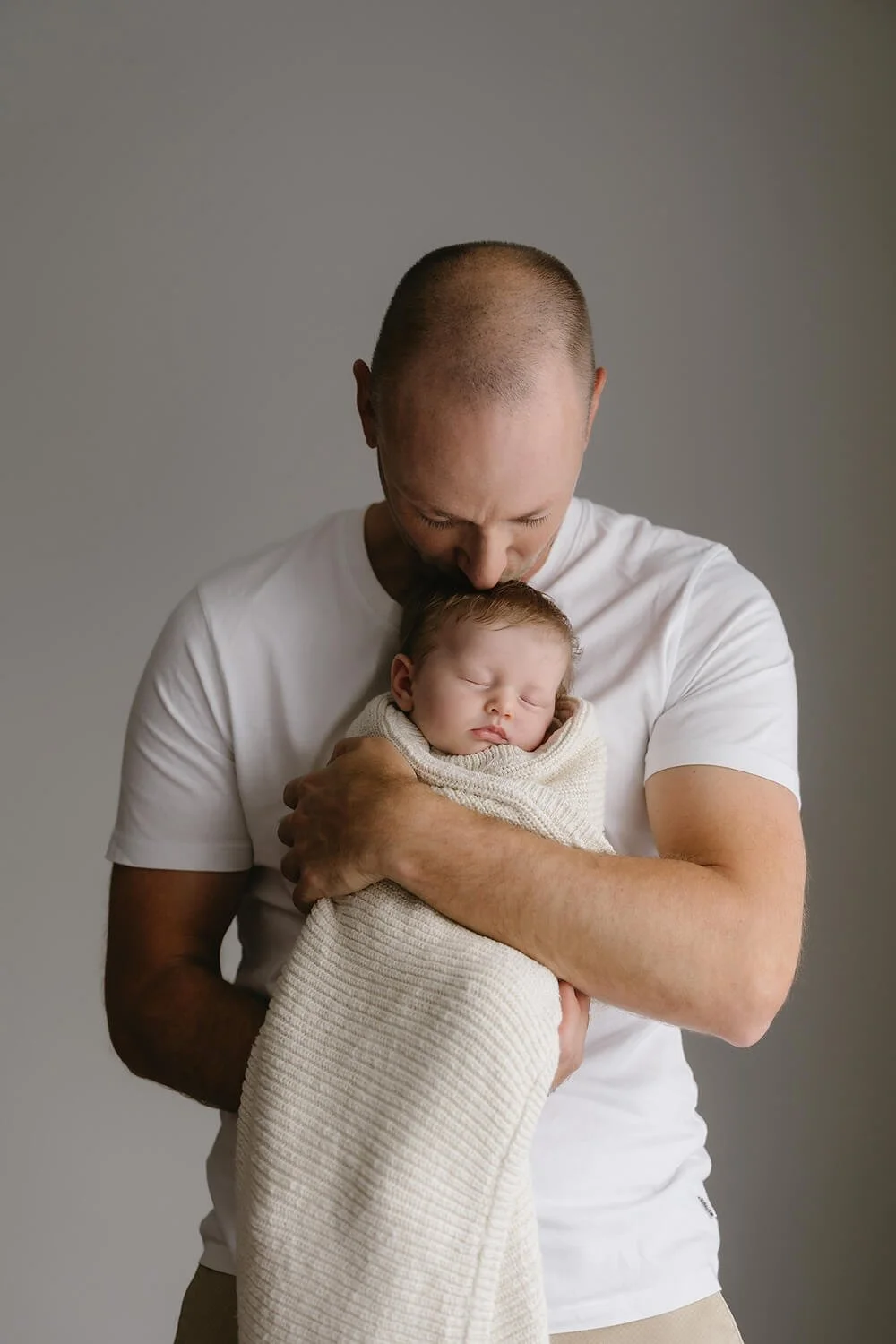 A father holding a sleeping baby wrapped in a blanket, with his head bowed down gently touching the baby's head.