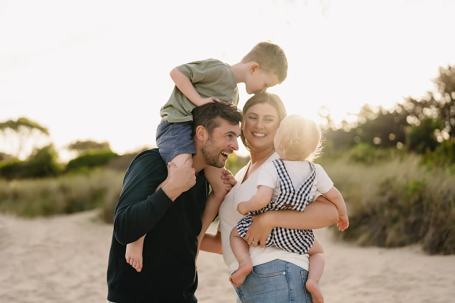 A happy family of four outdoors at sunset, with the father carrying a young boy on his shoulders, the mother holding a toddler boy, and their son leaning in to kiss the mother.