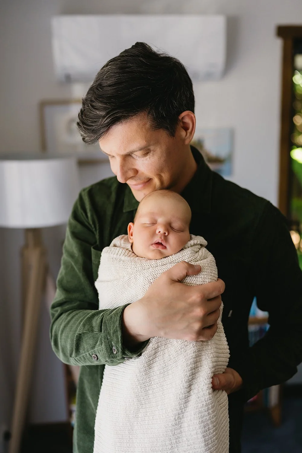A man holding a sleeping baby wrapped in a beige blanket, smiling softly while looking down at the child in a cozy indoor setting.