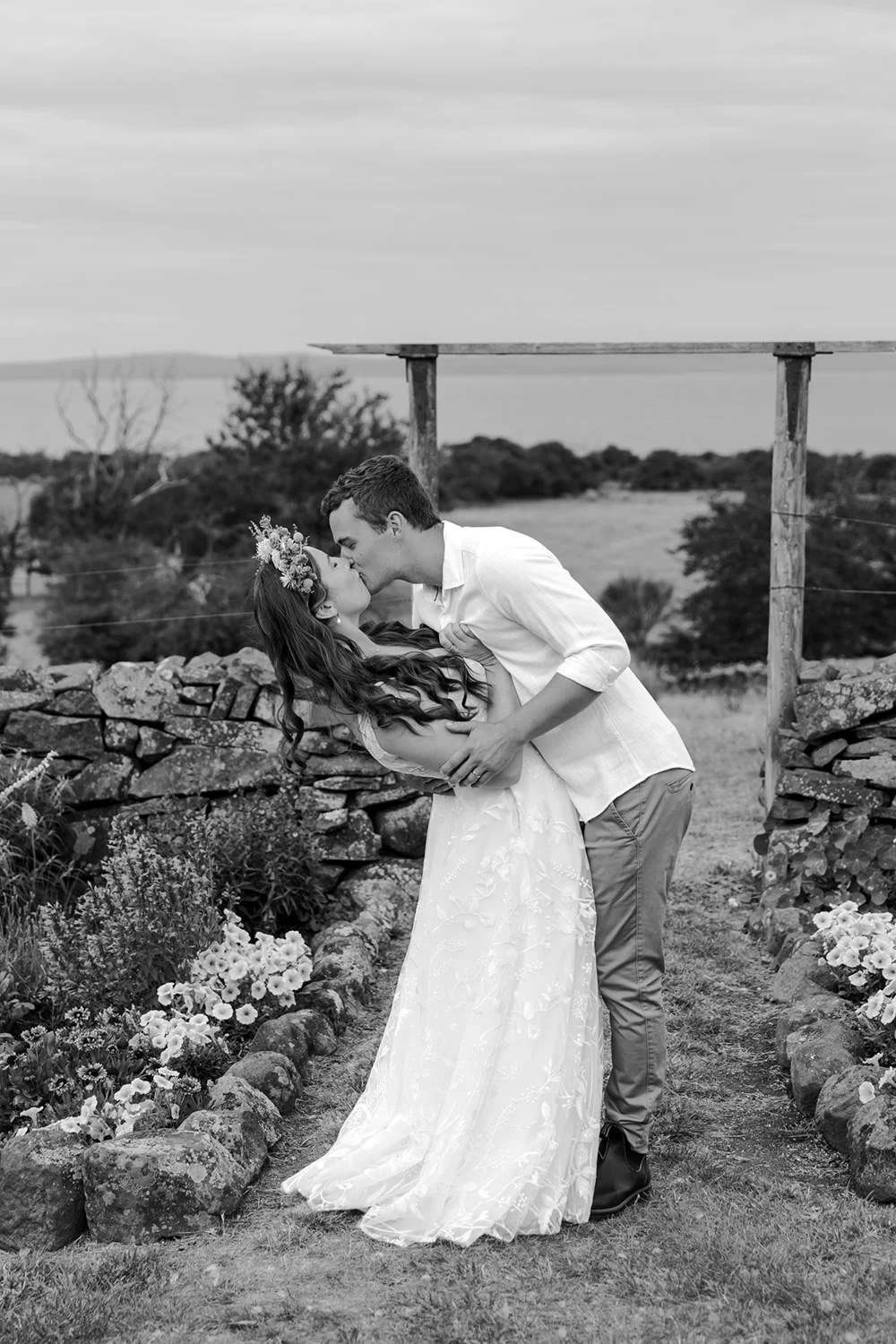 A couple kissing outdoors, the woman in a wedding dress and floral crown, the man in a white shirt, in a rural setting with a stone wall and flowers.