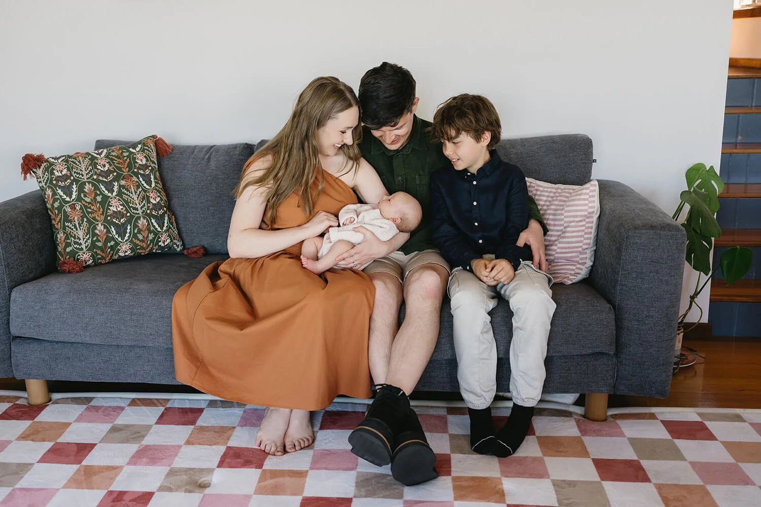 A family of four sitting on a gray sofa with wooden legs, smiling and looking at a newborn baby. The mother, wearing a brown dress, holds the baby, while the father, wearing a green shirt and beige shorts, sits between her and a boy in a navy shirt a