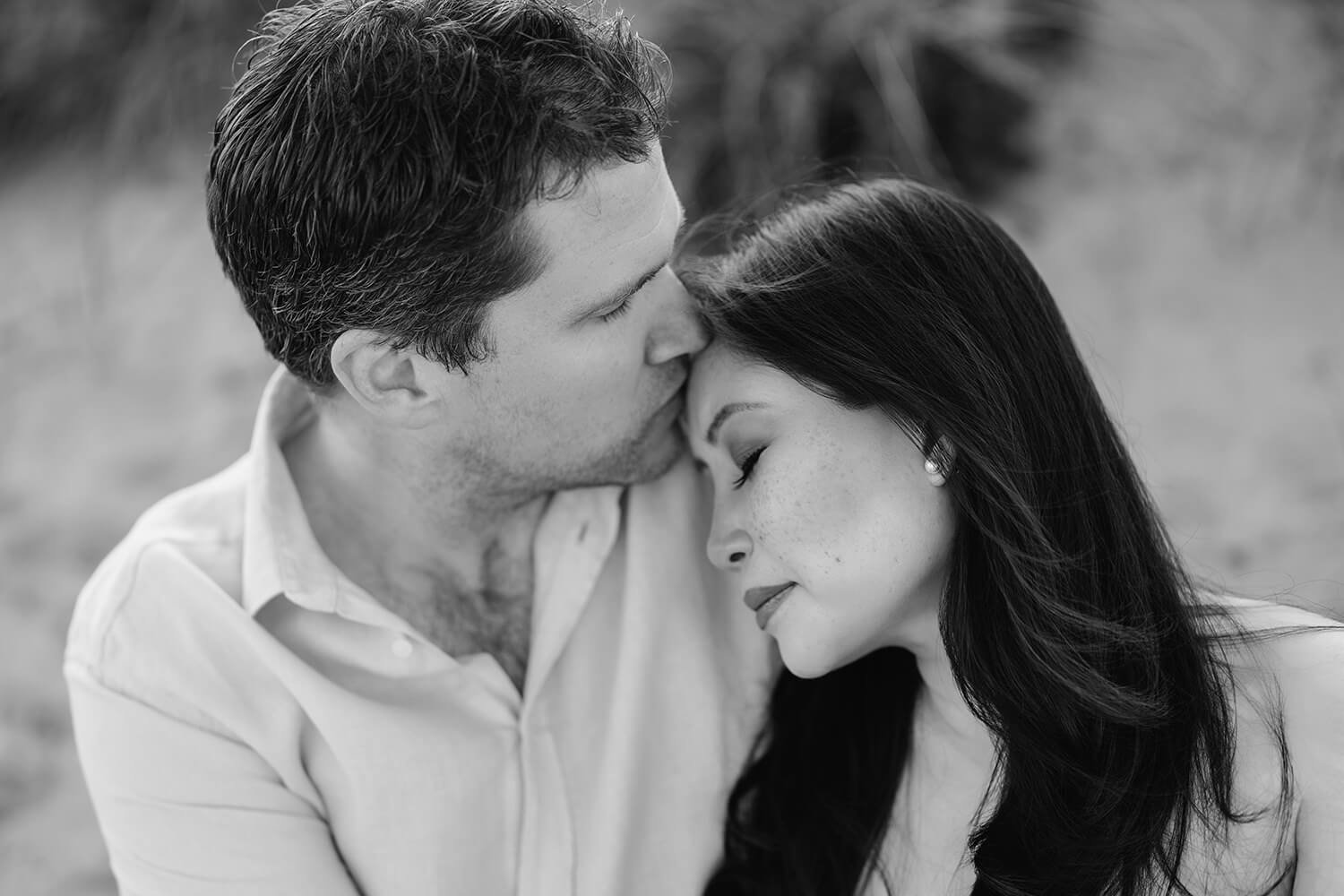 Black and white photo of a man kissing a woman on the forehead with her eyes closed, both with relaxed expressions, outdoors.