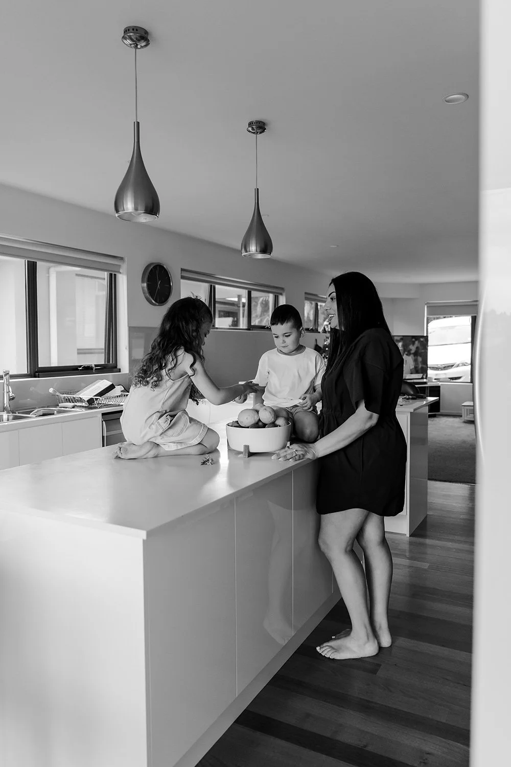A woman and two children in a kitchen, with one child sitting on the counter and the other standing, surrounded by fruit in a bowl, and a television and windows in the background.