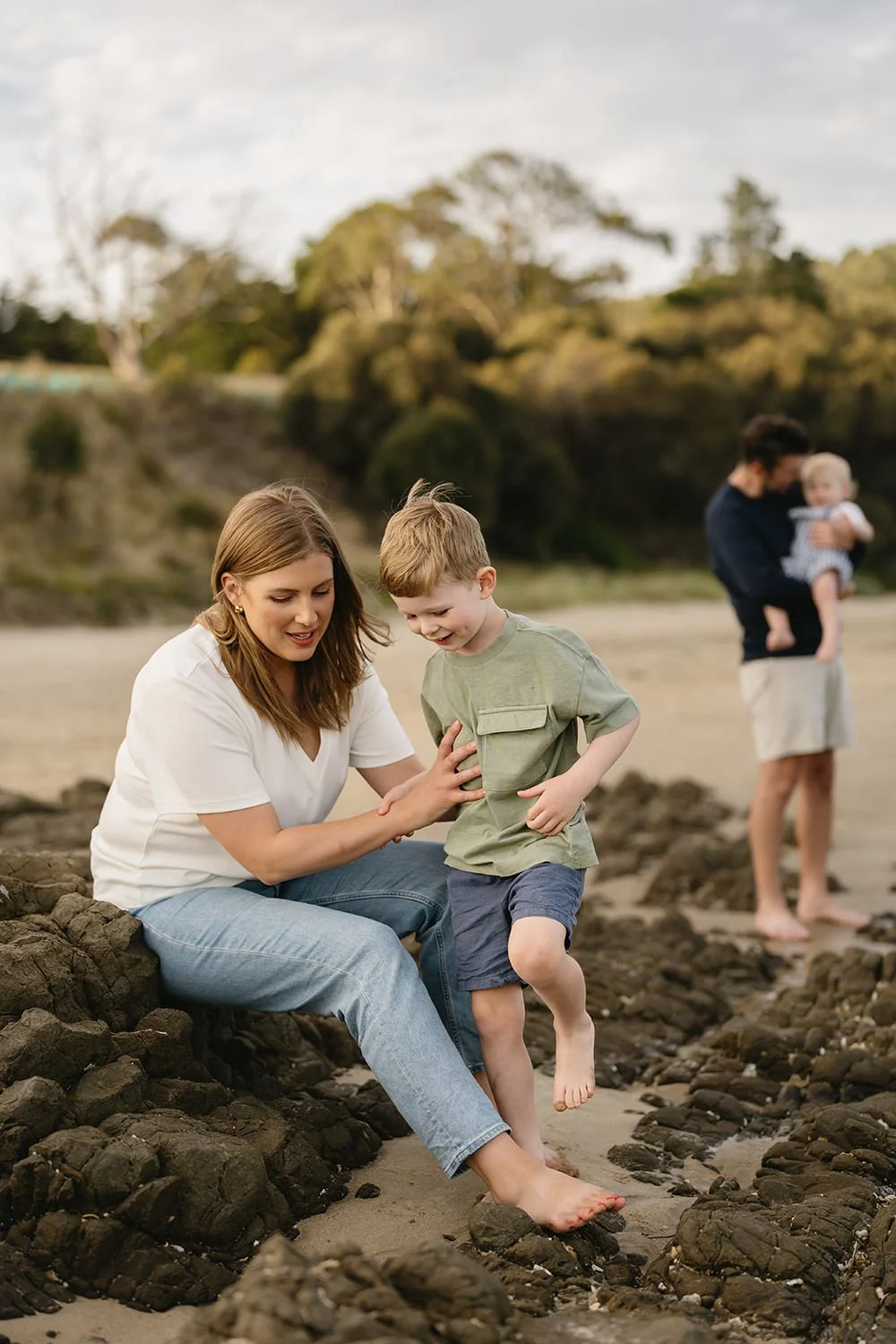 A woman and a young boy playing on rocks at the beach, with a couple and a child in the background.