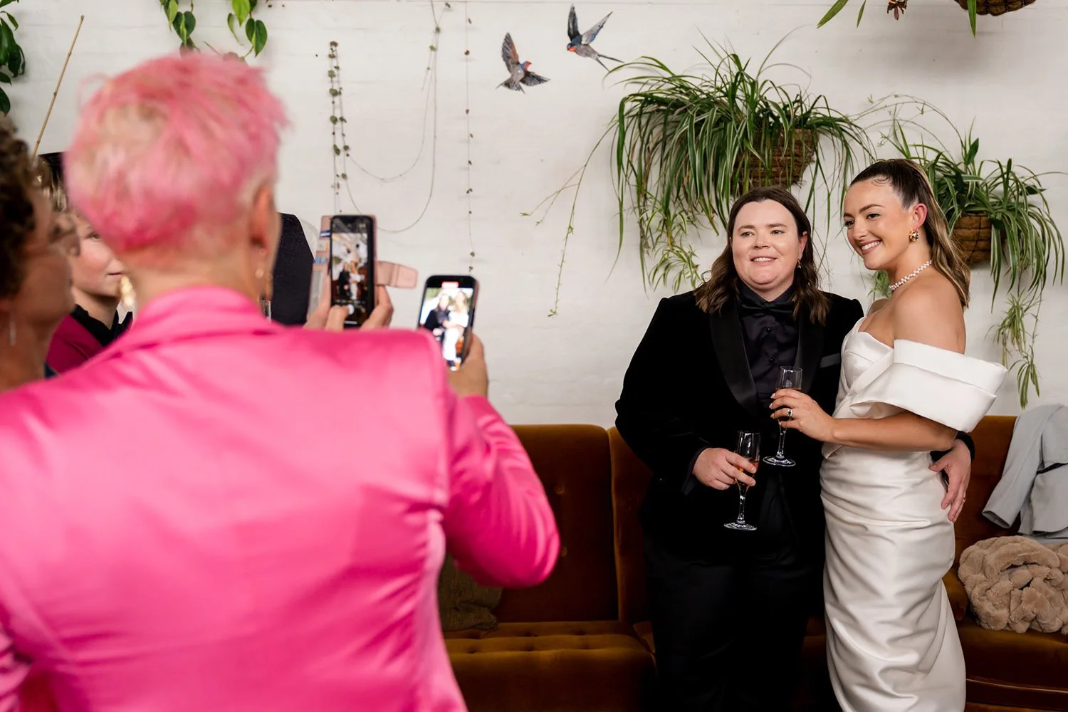 Two women, one in a black tuxedo and the other in a white off-shoulder dress, posing for photographs with guests taking pictures at a celebration in a decorated indoor venue with hanging plants and wall art.