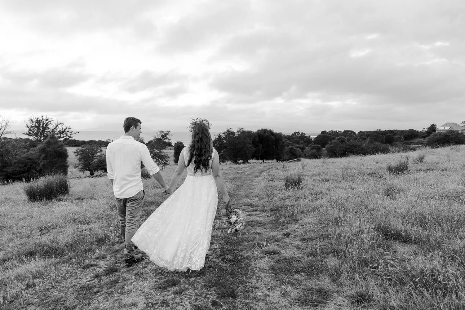 A black and white photo of a couple walking hand in hand through a grassy field, with the woman in a long dress and the man in a white shirt and pants, holding a bouquet of flowers.