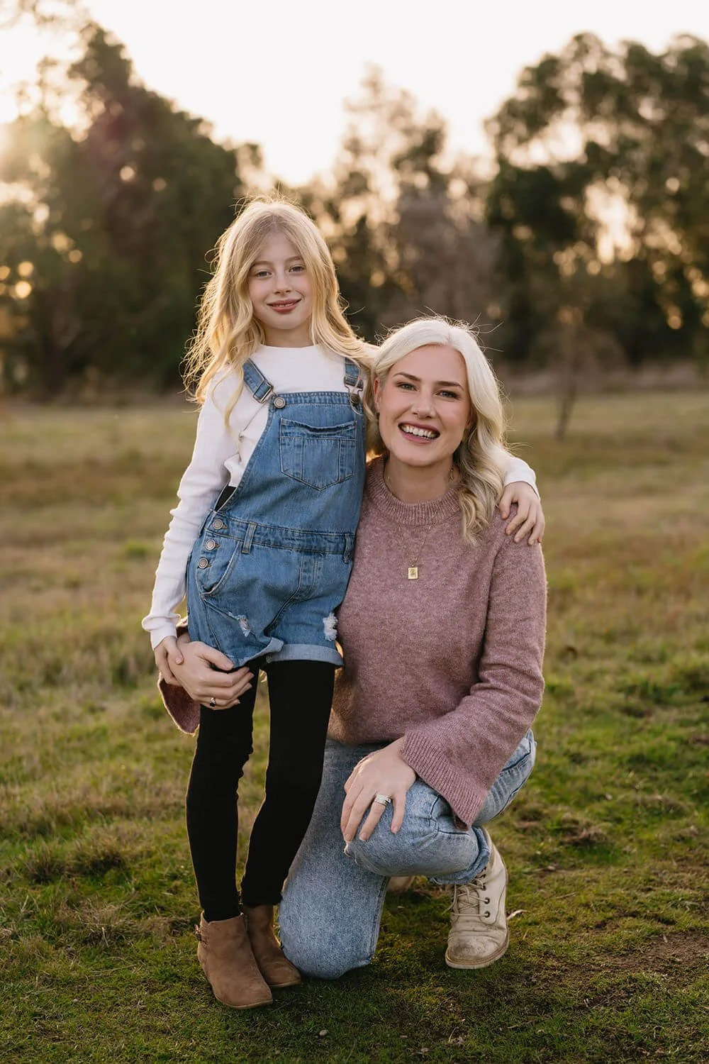 A woman kneeling on one knee with a little girl standing beside her outdoors during sunset, both smiling, in a grassy field with trees in the background.