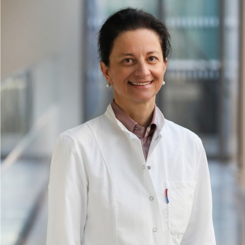 A smiling woman in a white lab coat standing in a modern medical or laboratory setting with glass windows.