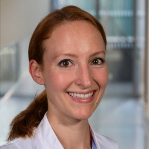 A woman with red hair tied back, smiling, wearing a white coat, in a professional indoor setting.