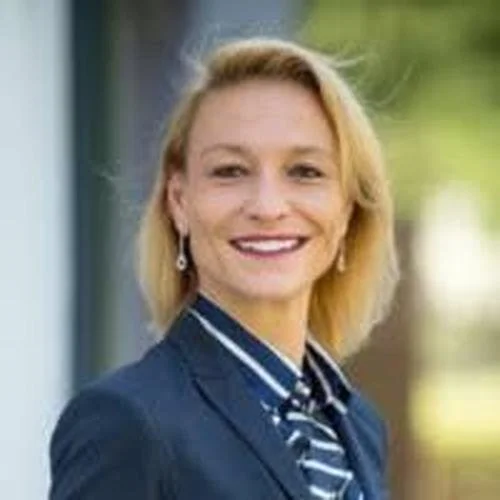 A smiling woman with blonde hair standing outdoors, wearing a navy blazer and striped shirt.
