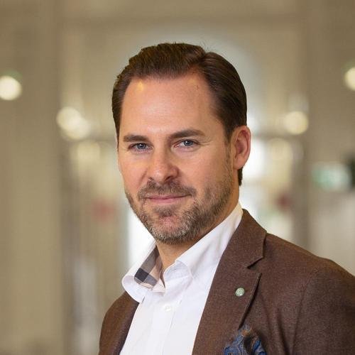 A man with short brown hair and a beard, wearing a brown suit jacket and a white shirt, standing in an indoor setting with blurred lights in the background.