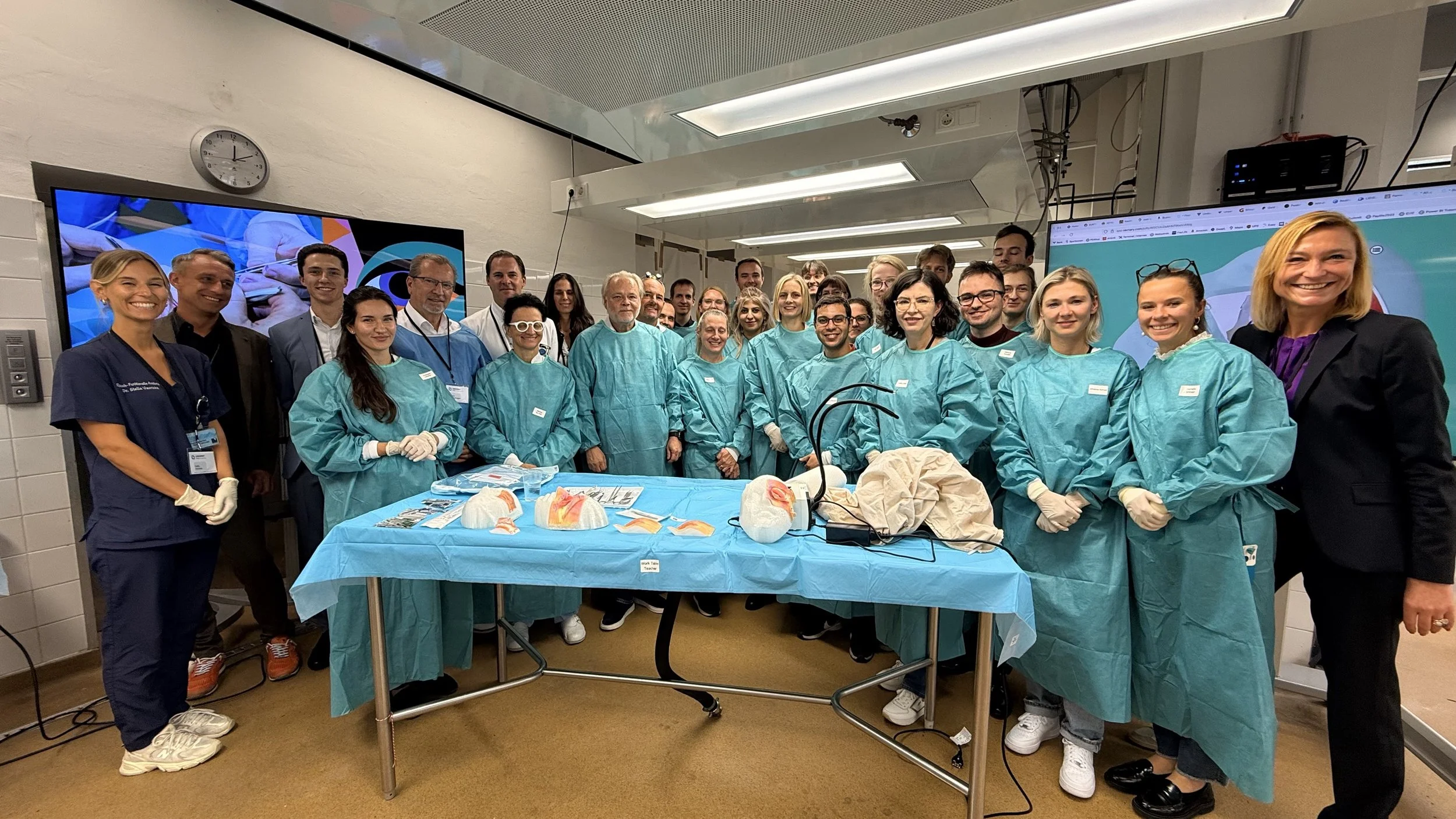 Group of medical professionals and staff in a hospital room, dressed in scrubs and lab coats, standing around a table with medical models and equipment, smiling for a photo.