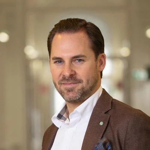 A man with short dark hair, light skin, and a beard wearing a brown blazer and white shirt in an indoor setting with blurred lights in the background.