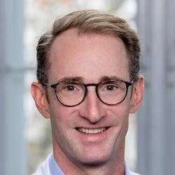 Close-up of a smiling man with glasses and short light brown hair, wearing a light blue shirt, against a blurred office background.