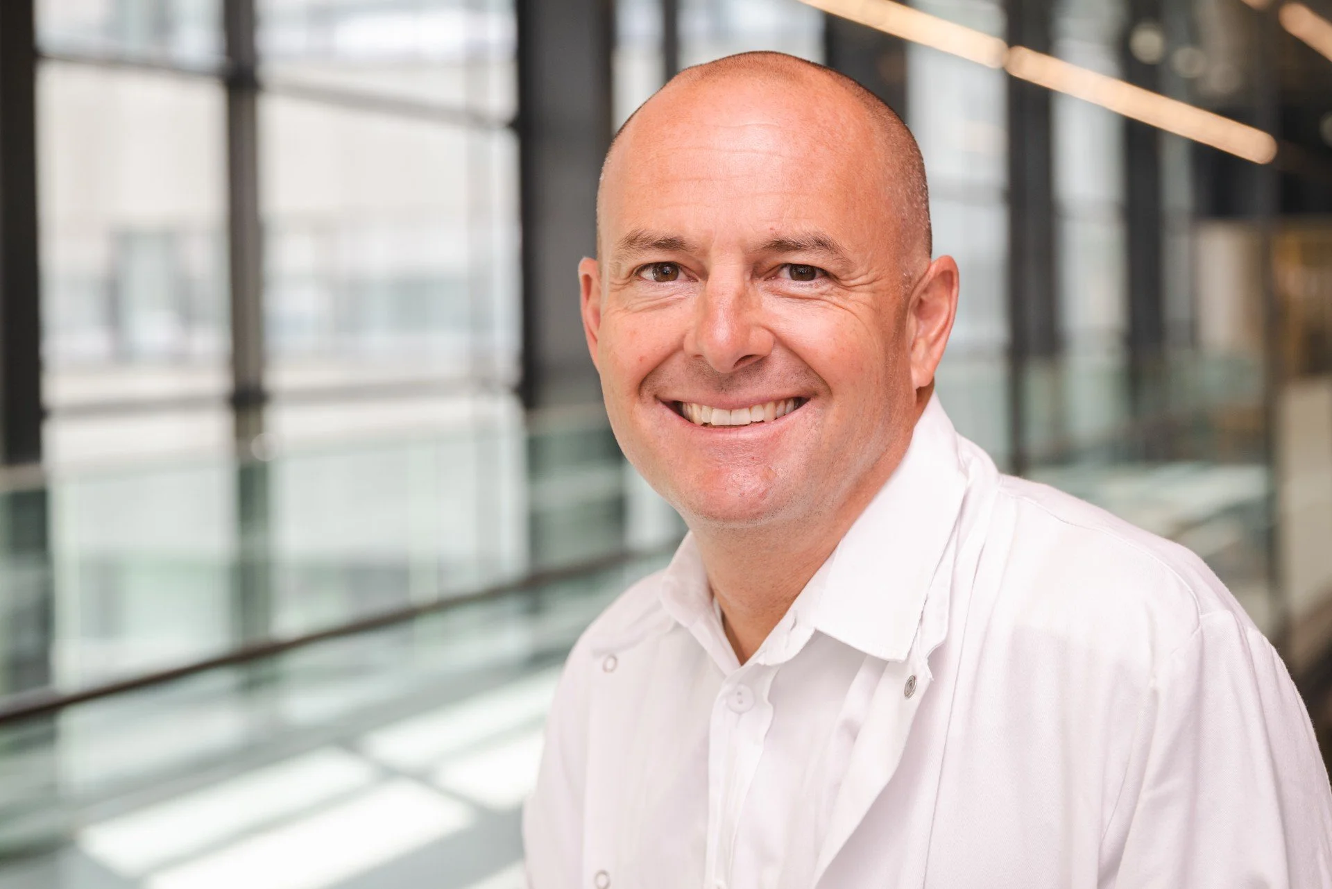 A smiling male doctor in a white lab coat and blue tie, standing in a medical or laboratory setting.