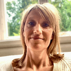 A woman with blonde hair smiling indoors near a window with trees outside.