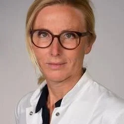 A woman with blonde hair wearing glasses and a white coat.