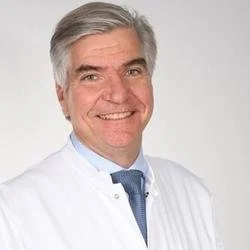 A smiling middle-aged man with gray hair in a white medical coat and blue tie, posing against a plain light background.