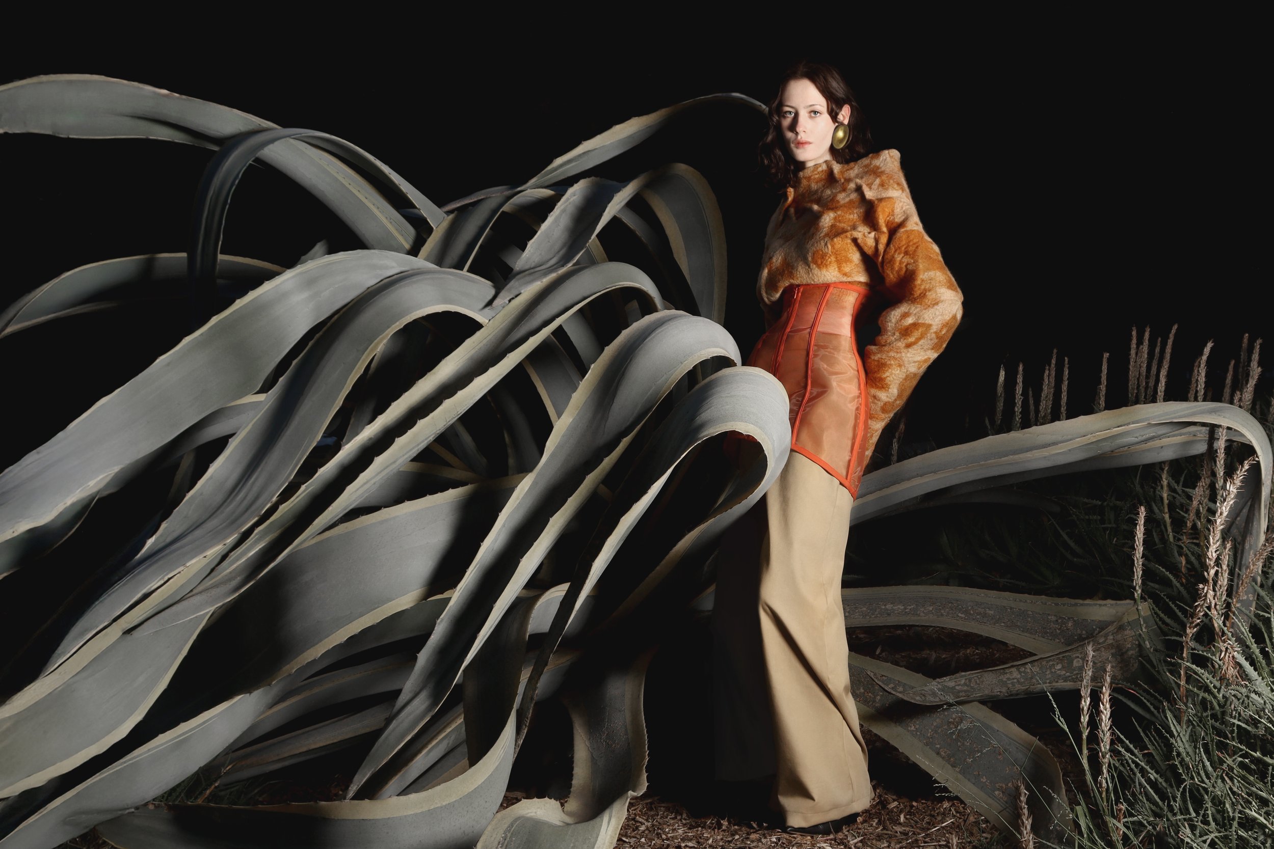 A woman in a fur coat, beige wide-leg pants, and large gold earrings stands beside oversized agave plant leaves against a dark background.