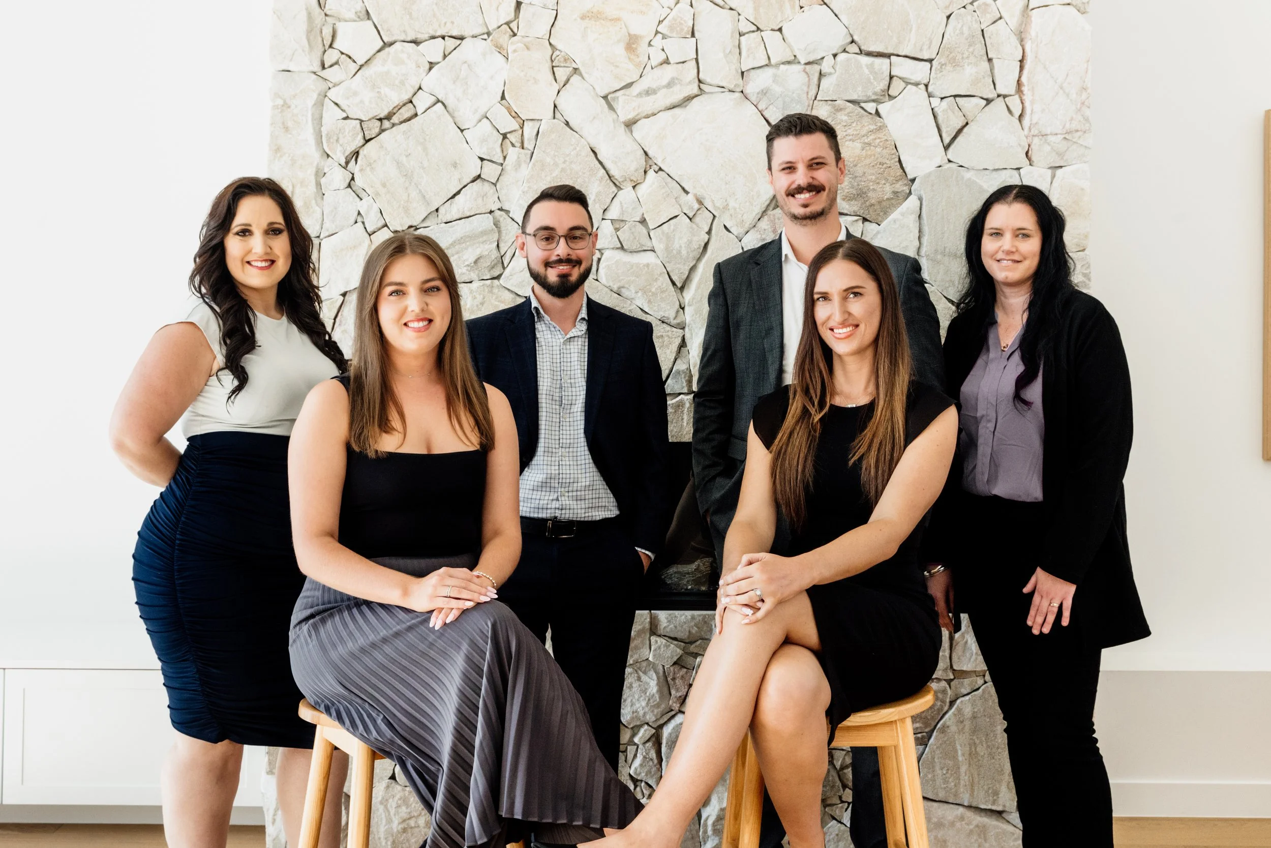 A group of six professionally dressed people posing together indoors in front of a stone wall, smiling.