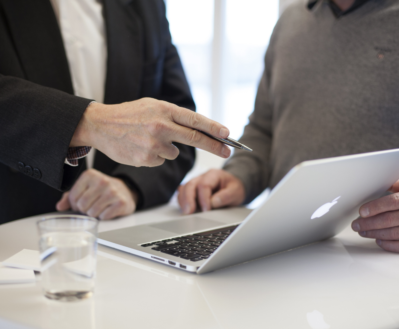 Two men in business attire discussing a document with a pen in front of a MacBook laptop and a glass of water on a white table.