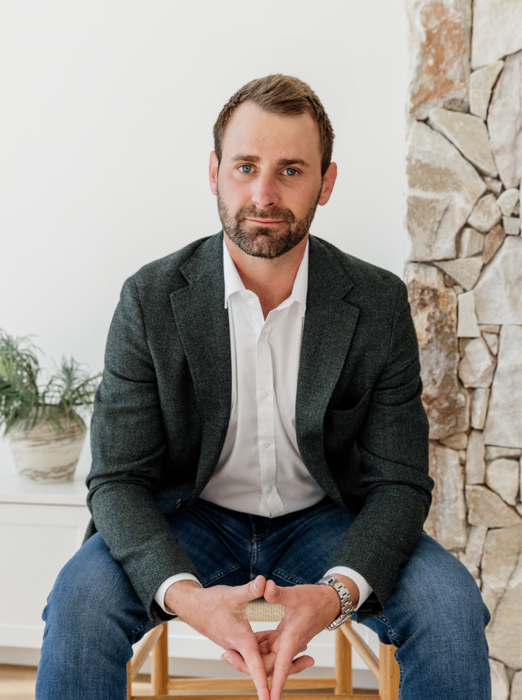 A man in a white shirt and dark blazer sitting on a wooden chair indoors, with a stone wall to his right and a white cabinet with potted plants in the background.