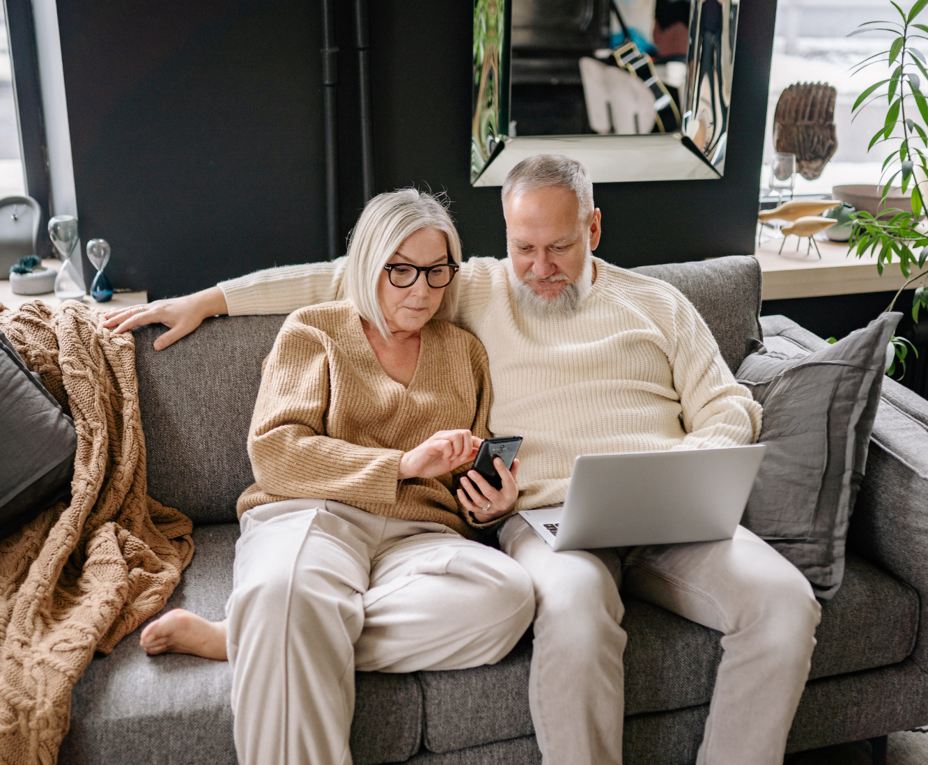 An elderly woman and man sitting on a gray couch, looking at a smartphone together. The woman wears glasses and a tan sweater, and the man wears a cream sweater. They are in a cozy living room with decorative items and a mirror in the background.