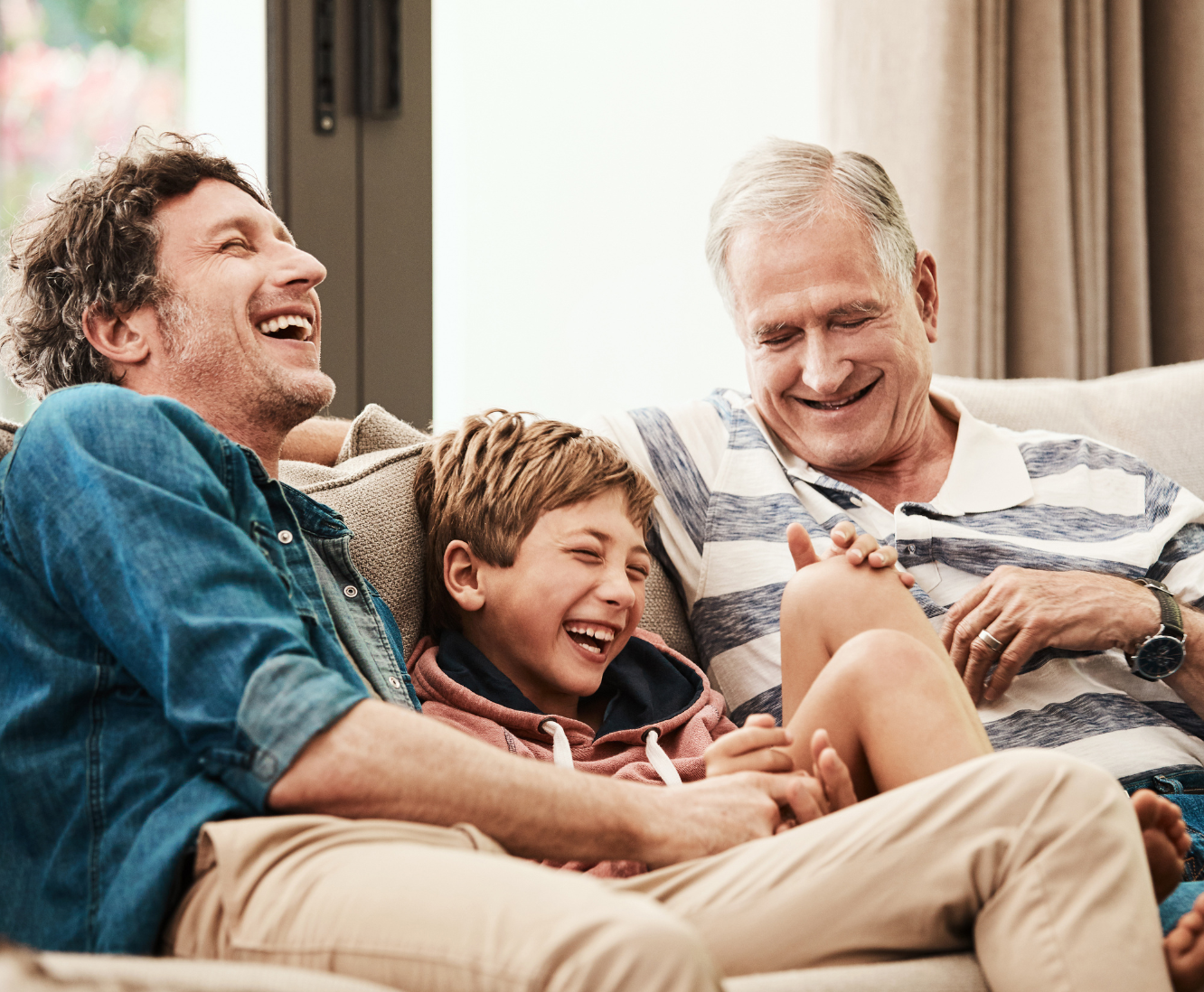 Three generations of men, a father, grandfather, and son, sitting on a couch, laughing and enjoying time together.