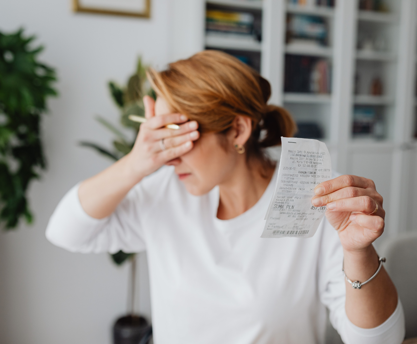 A woman with red hair holding a receipt in one hand and pressing her forehead with the other, appearing stressed or worried, indoors with bookshelves and a plant in the background.