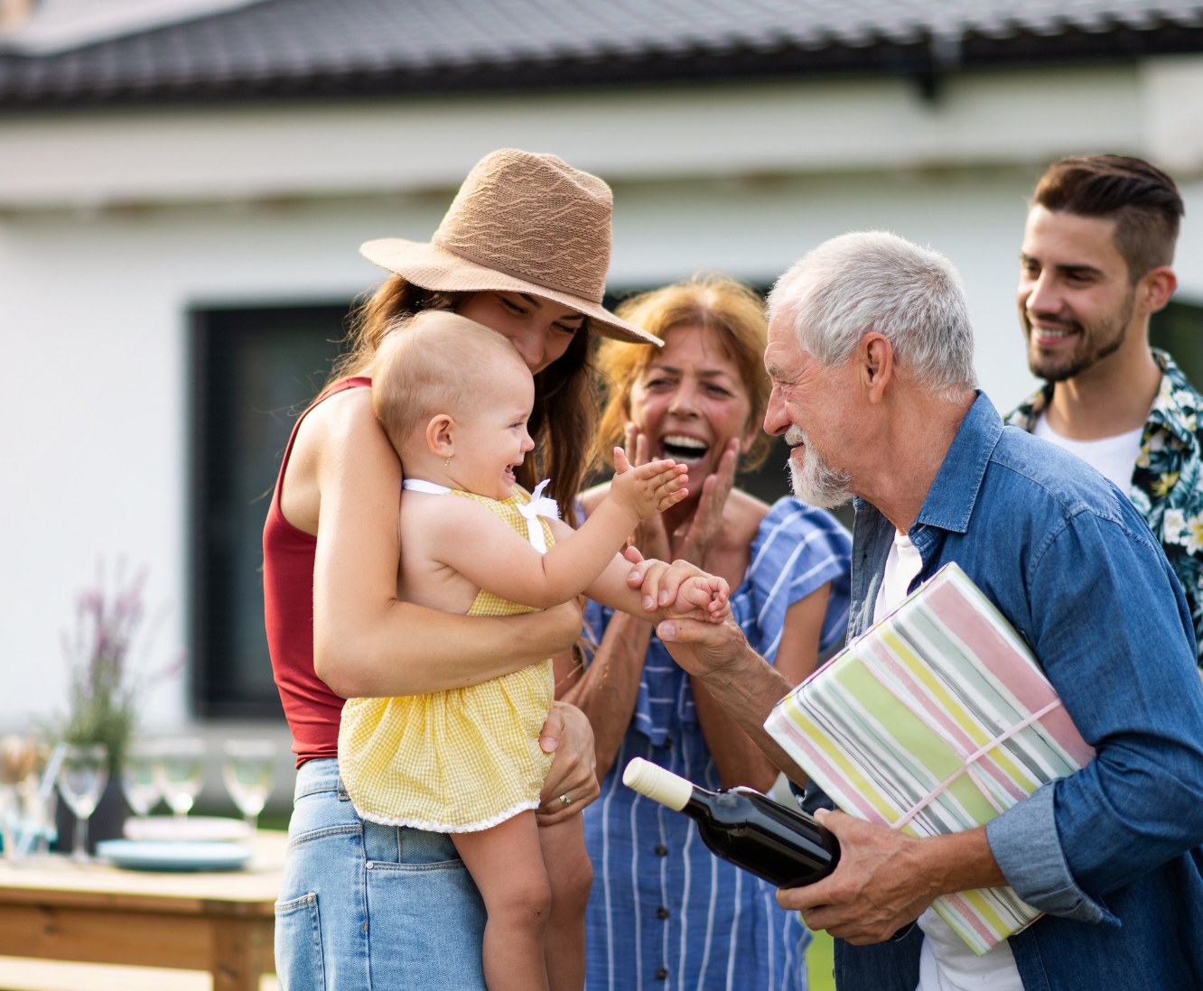A family gathering outdoors with two women, an older man, a young man, and a small girl smiling and interacting. The older man is holding a wrapped gift and a bottle of wine, and the little girl is reaching out to shake the man's hand.