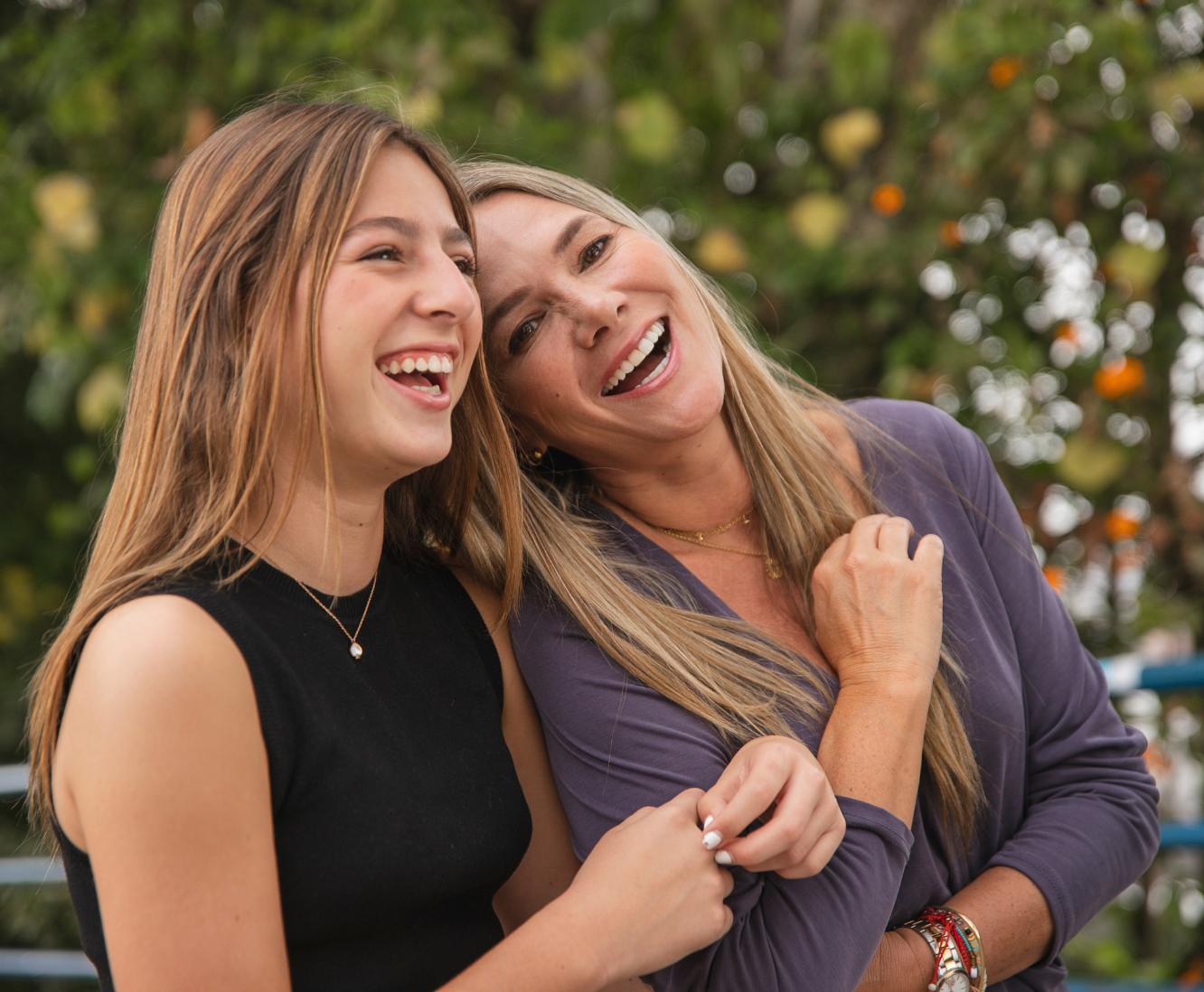Two women smiling and laughing outdoors with greenery and orange flowers in the background