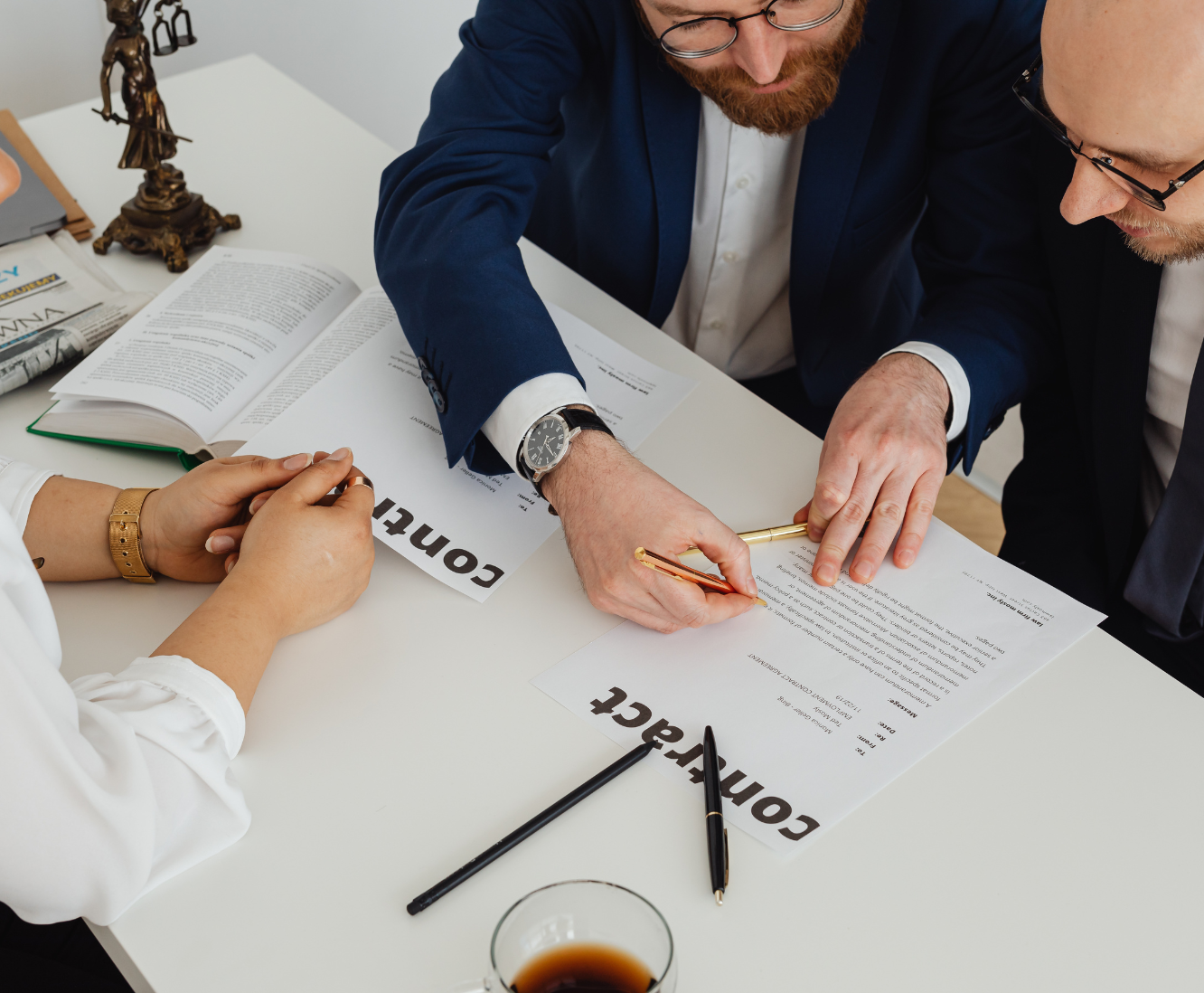 Two men in suits signing a contract while a woman watches, with documents, pens, and a newspaper on the table.