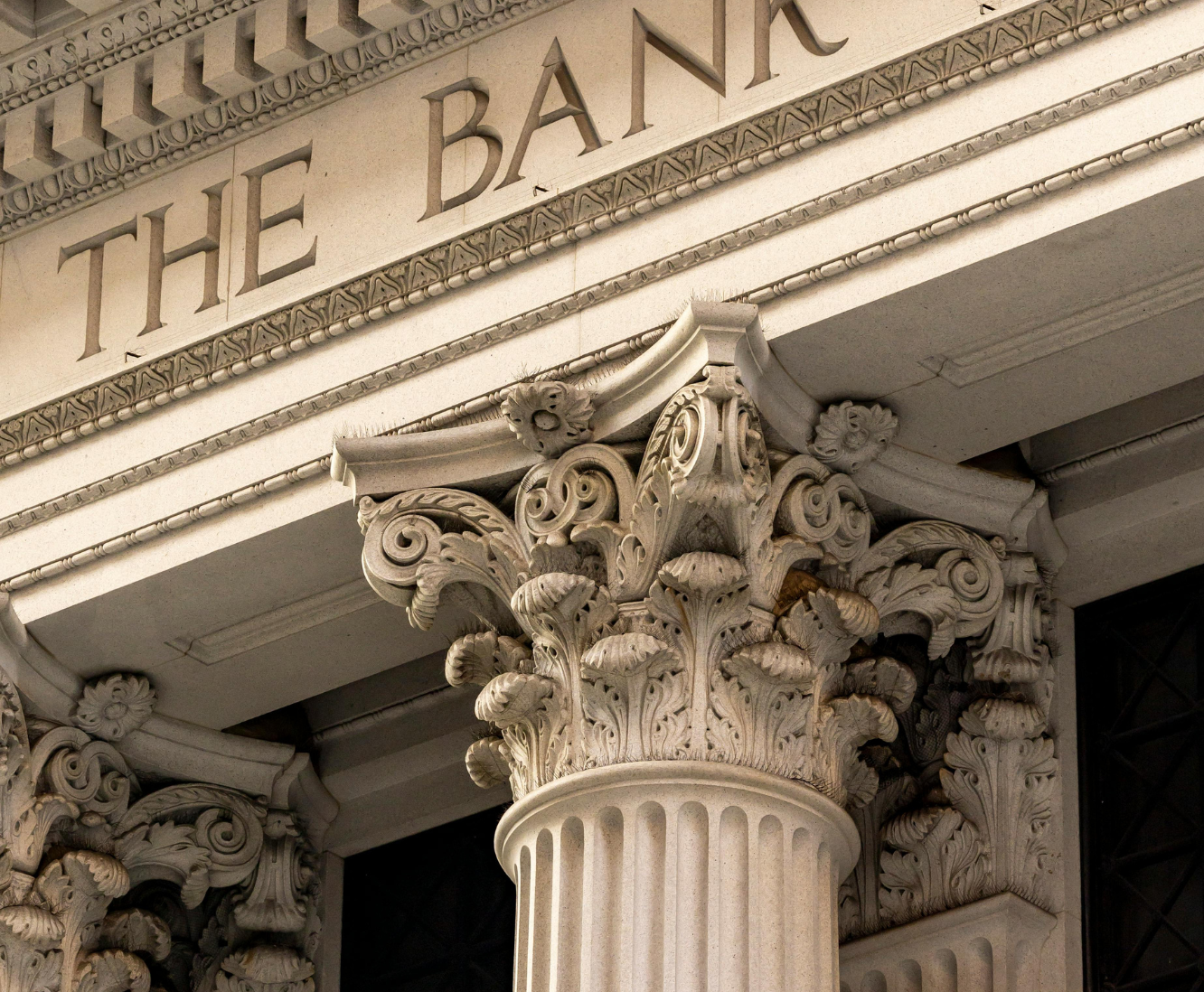 Close-up of the ornate corinthian column capital and the front facade of a bank building with the partially visible inscription 'THE BANK'