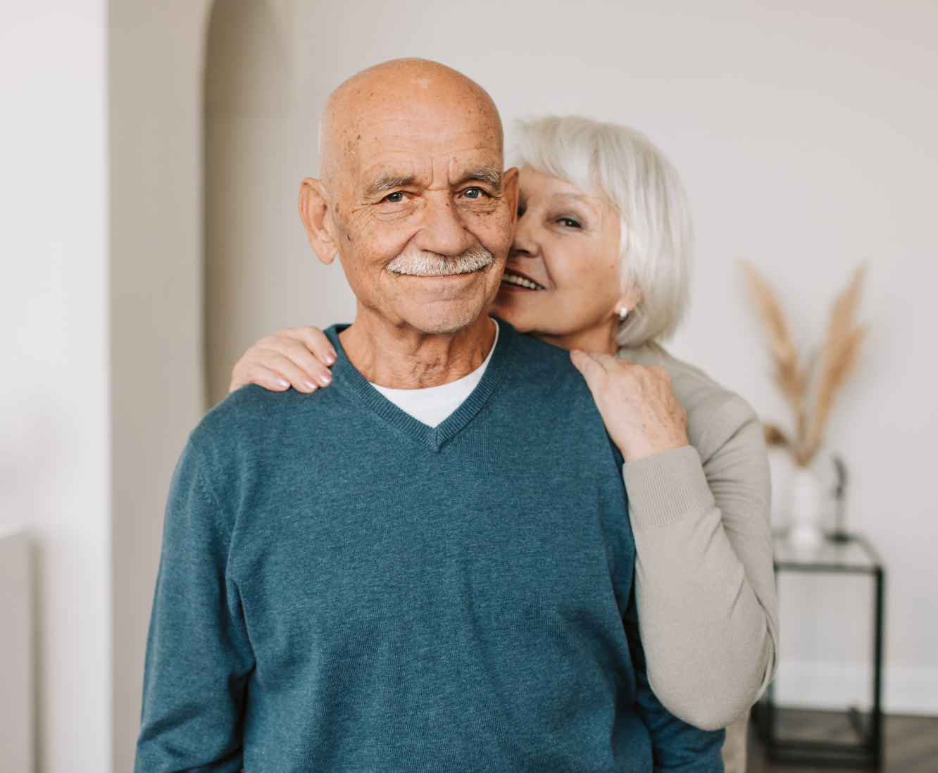 An elderly couple smiling and hugging in a home setting.