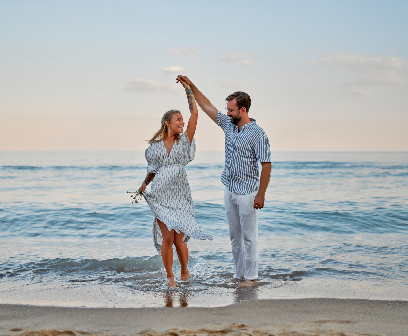 A smiling couple dancing in the shallow waves at the beach during sunset, with the woman holding a small bouquet and the man raising his hand.