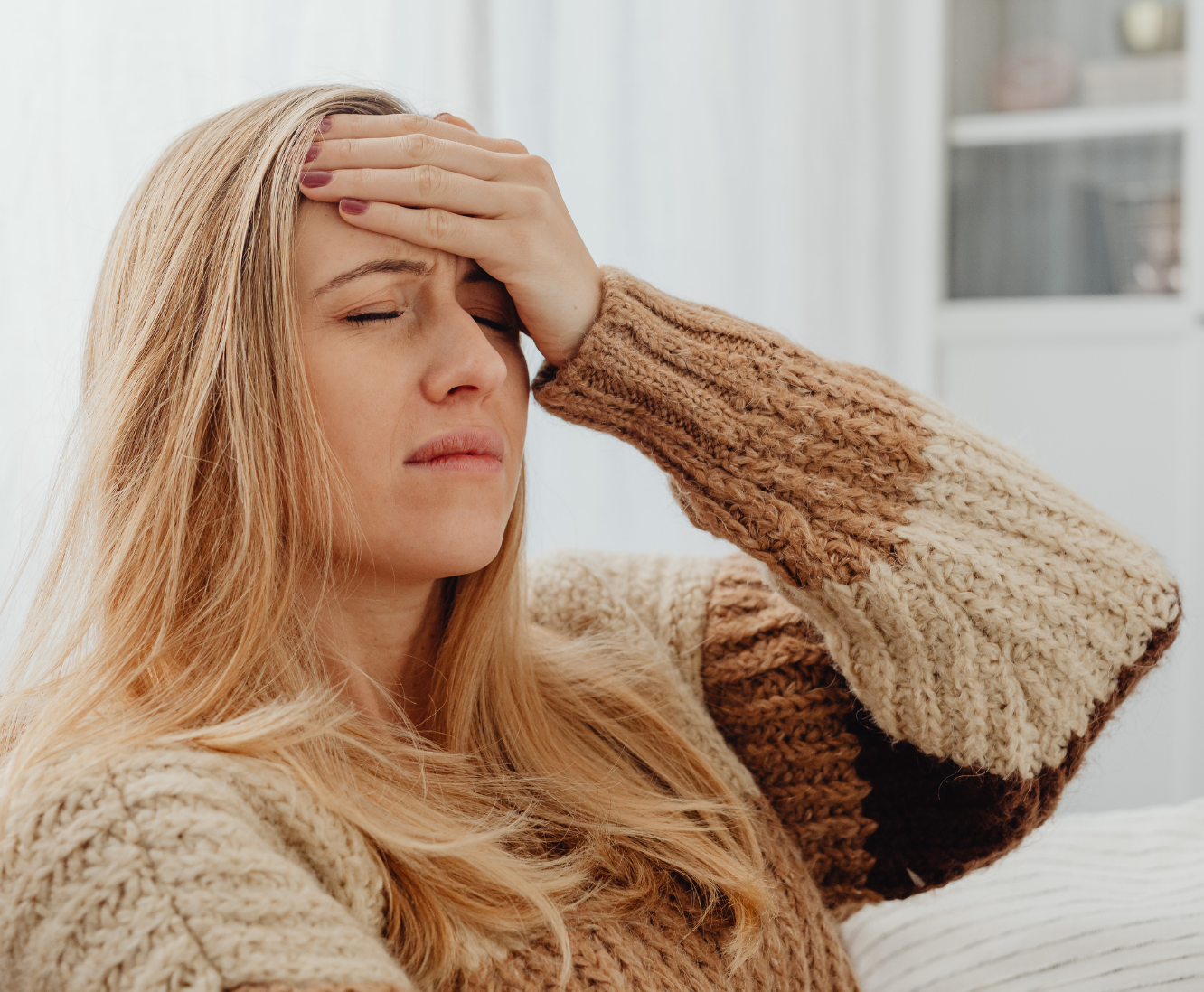 Young woman with long blonde hair in a cozy brown and beige sweater holding her forehead with her hand, appearing to have a headache indoors.