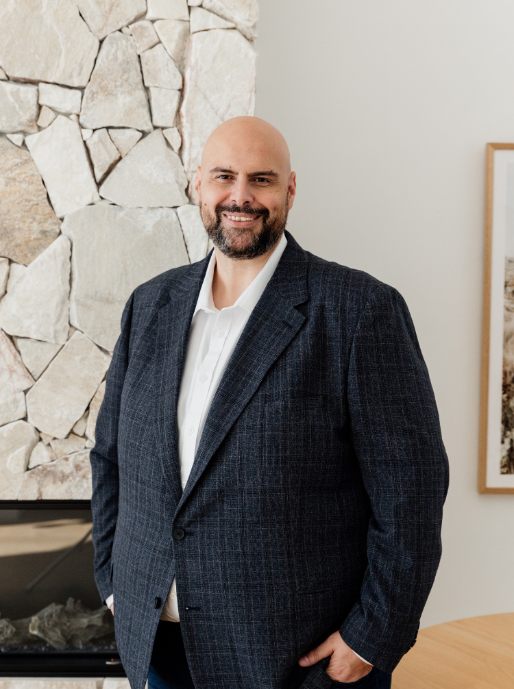 A man with a bald head and beard wearing a dark plaid blazer and white shirt, smiling while standing indoors near a stone wall and framed picture.