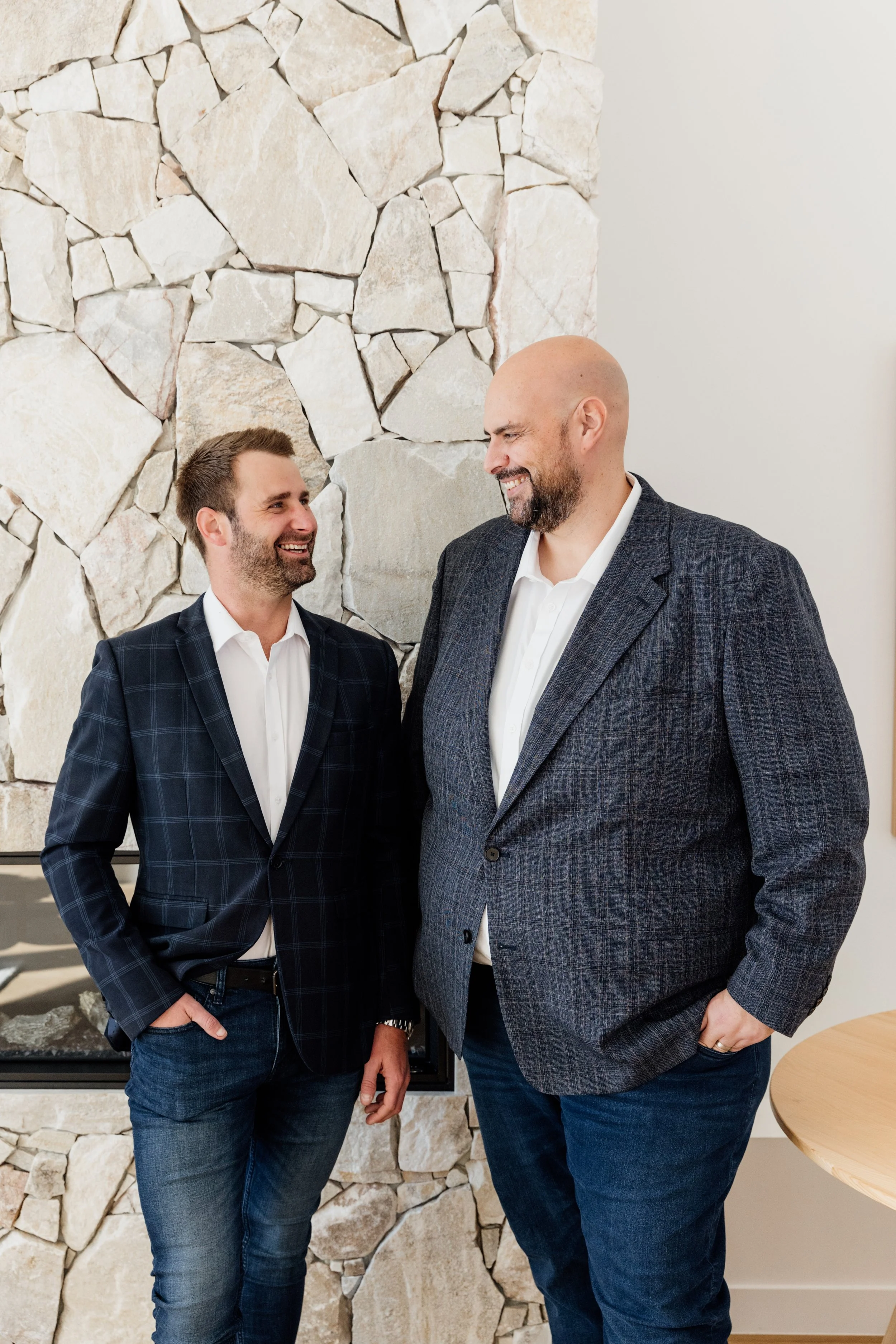 Two men in business casual attire smiling at each other indoors near a stone fireplace.