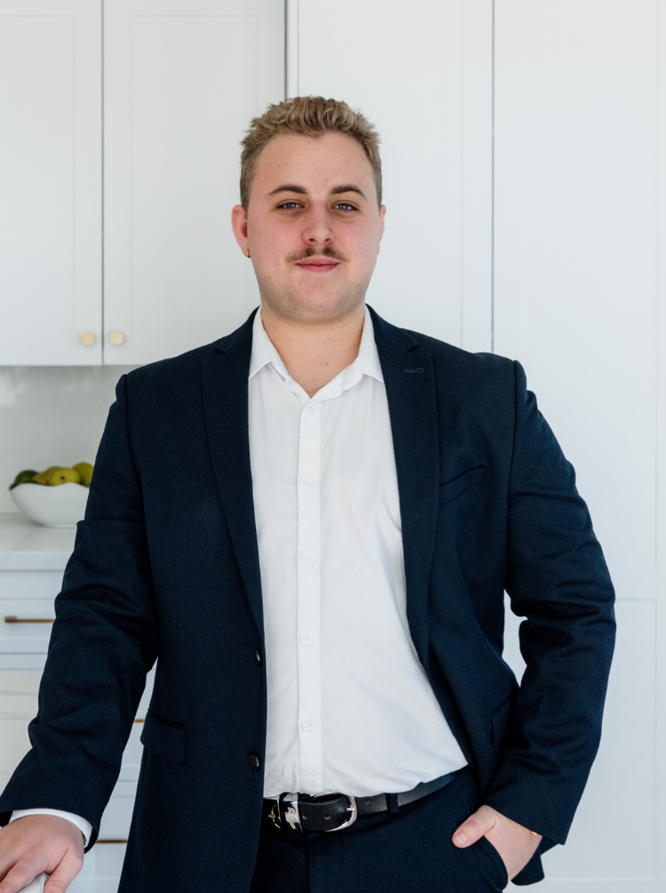 A young man with short blond hair, a mustache, and light skin, wearing a dark blue blazer and a white shirt, standing in a modern kitchen with white cabinets and a bowl of apples on the counter.