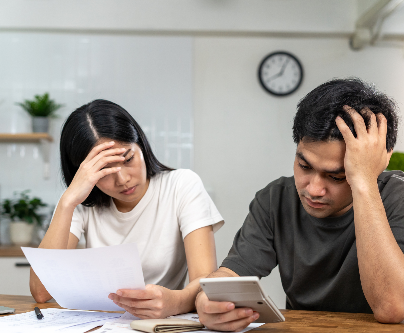 Two stressed people, a woman and a man, sitting at a table with documents and a smartphone, in a kitchen or office setting.
