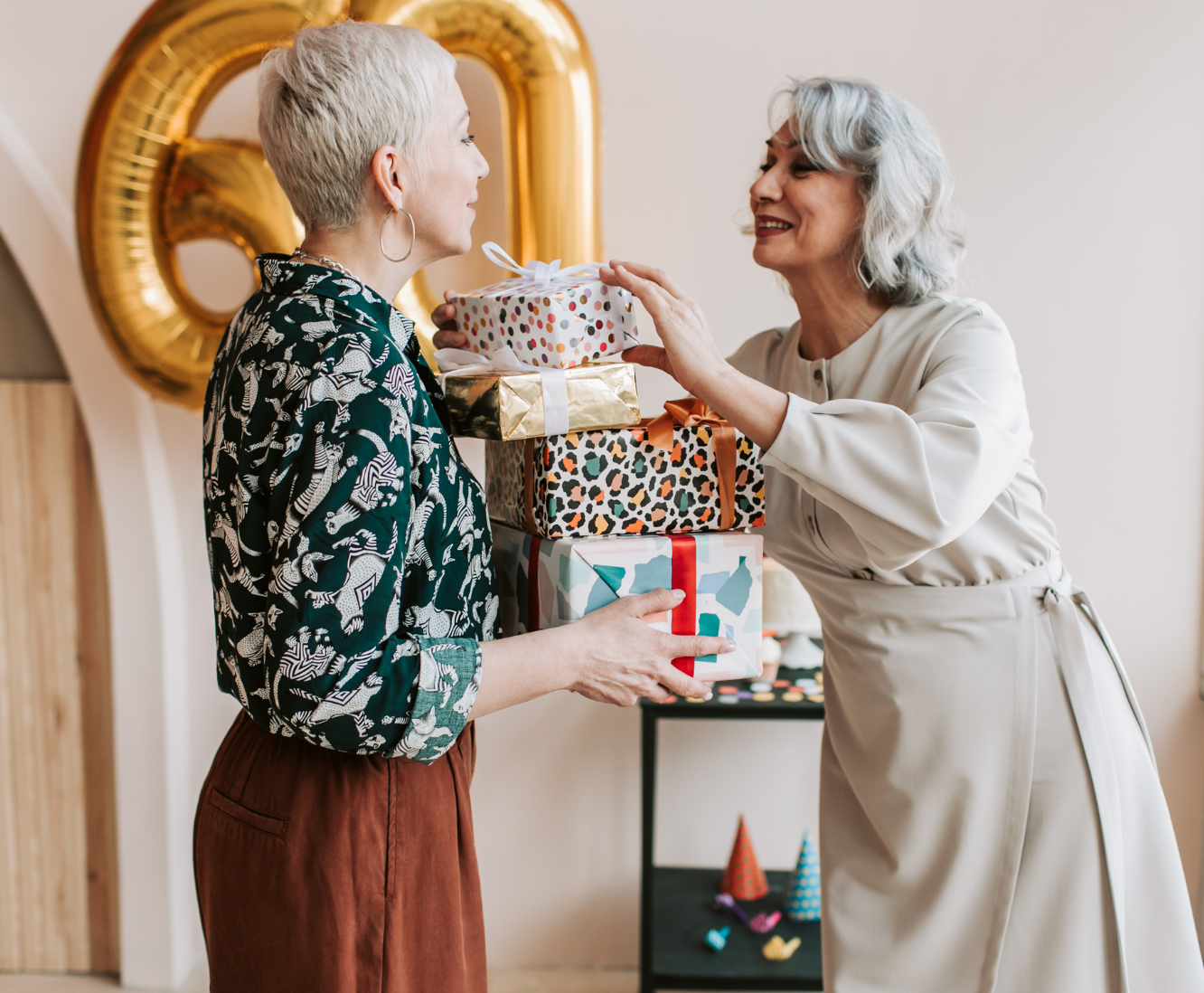 Two women exchanging gifts at a celebration, one woman handing a stack of wrapped presents to the other. Gold balloon in the background.
