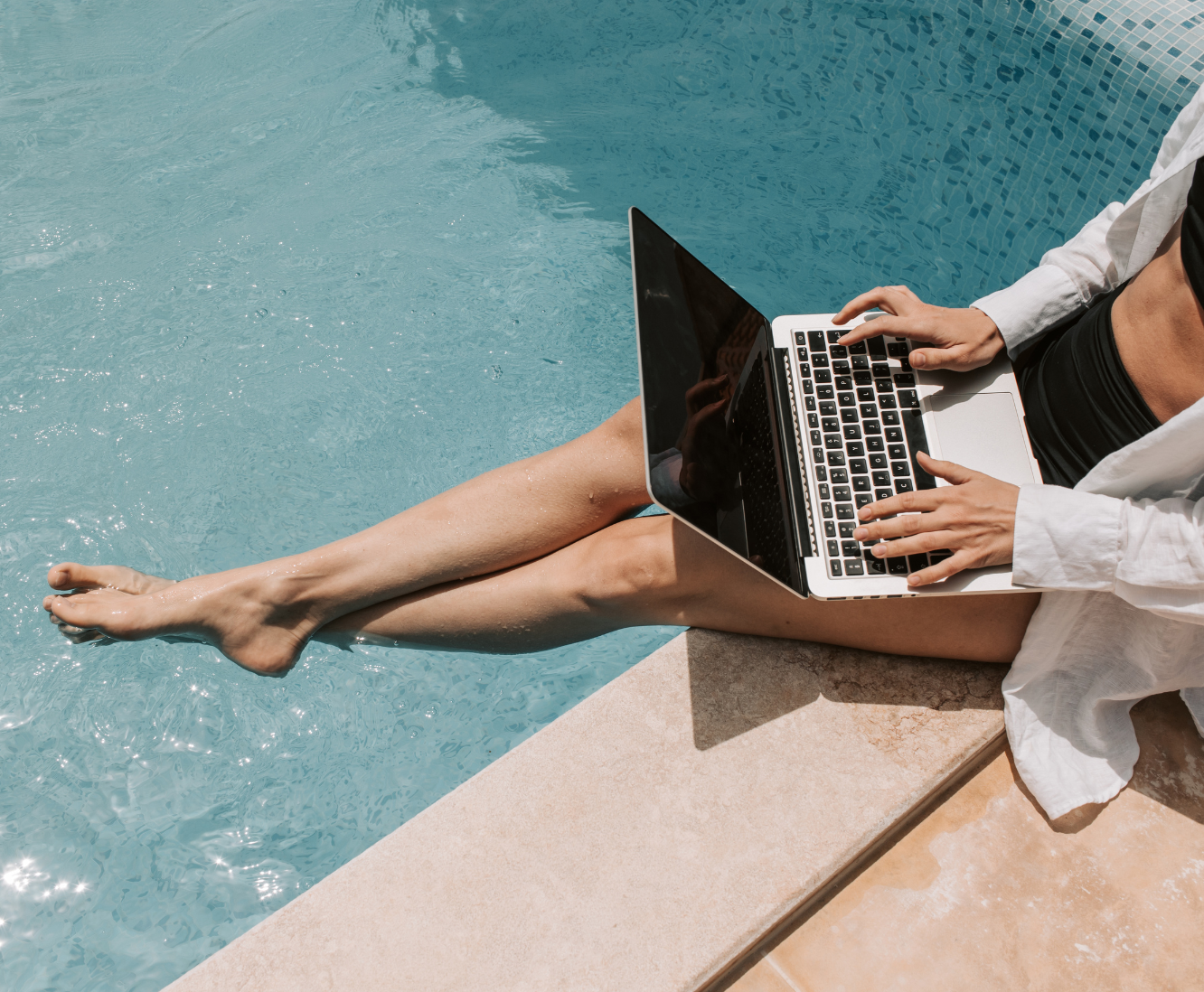 Person sitting at the edge of a swimming pool working on a laptop, with their legs in the water.