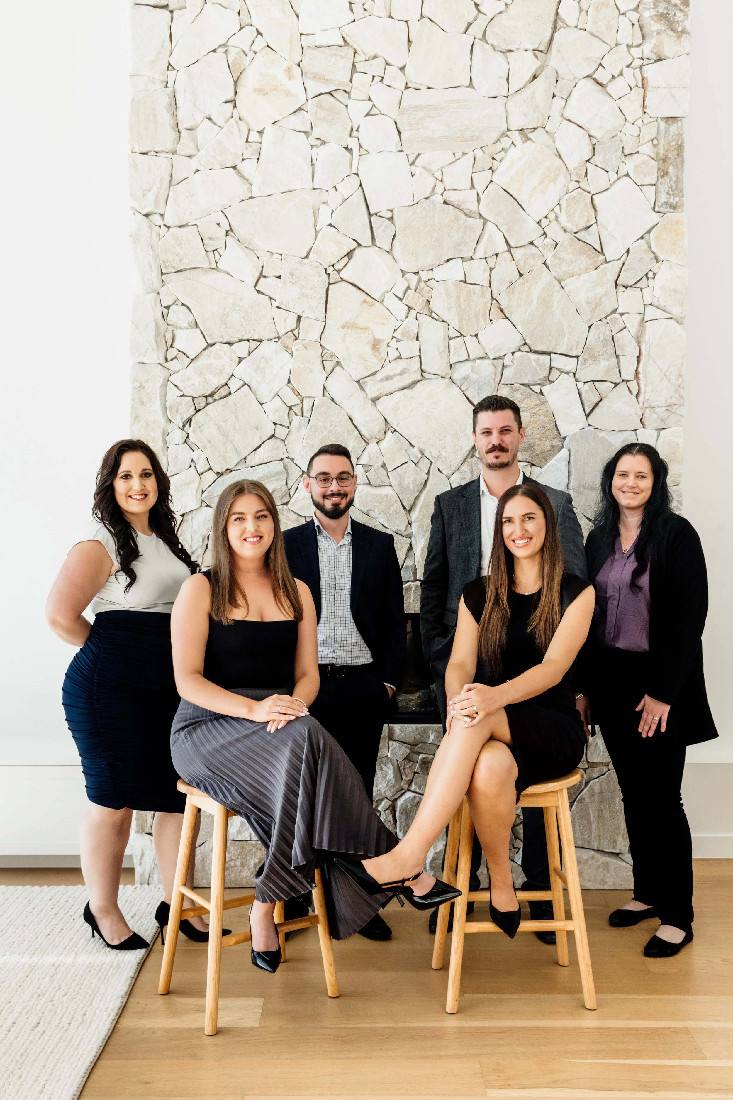 Group of six people posing for a photo in front of a stone fireplace, with two seated women in the front and four standing individuals behind them.