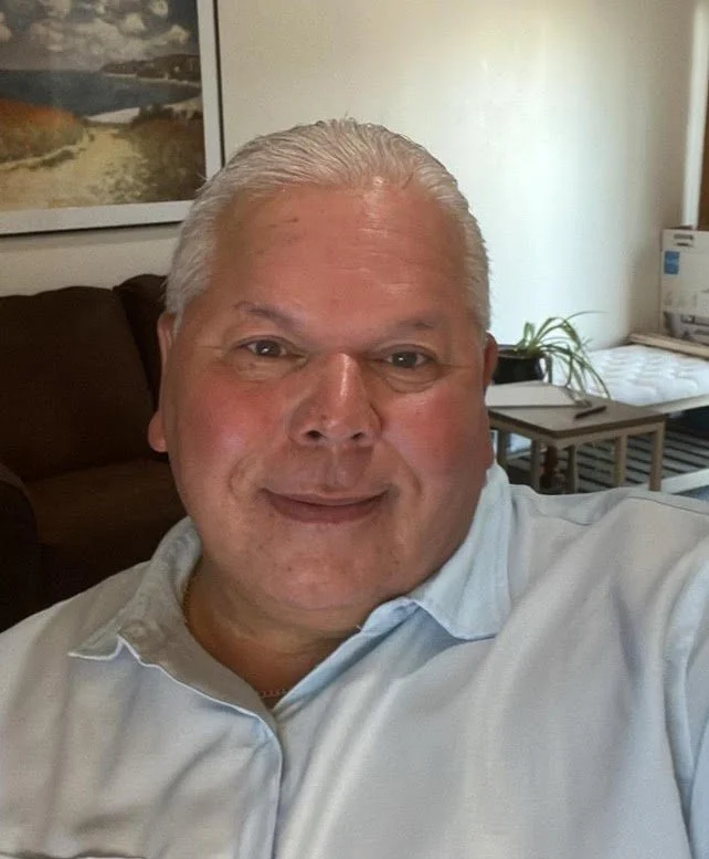 A middle-aged man with short white hair wearing a light blue shirt, smiling at the camera in a cozy indoor setting with artwork on the wall behind him and a small table with plants and boxes.